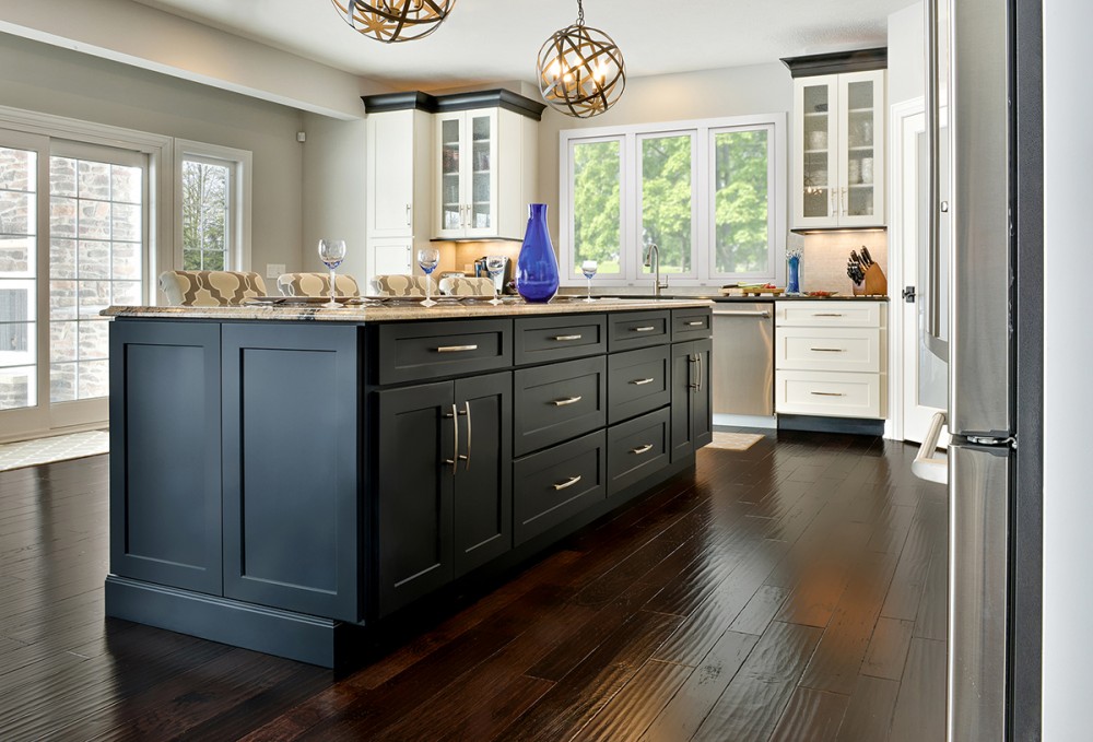 Open kitchen design in modern transitional style has large black island cabinets, black crown molding on white Shaker perimeter cabinets, and distressed hardwood flooring.